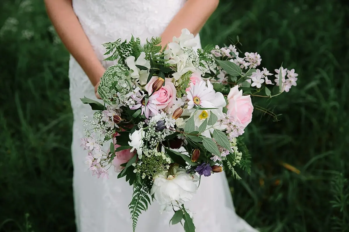 Bride in lace holding a wild bouquet of pink roses, ferns and white flowers against long grass at Sulgrave Manor
