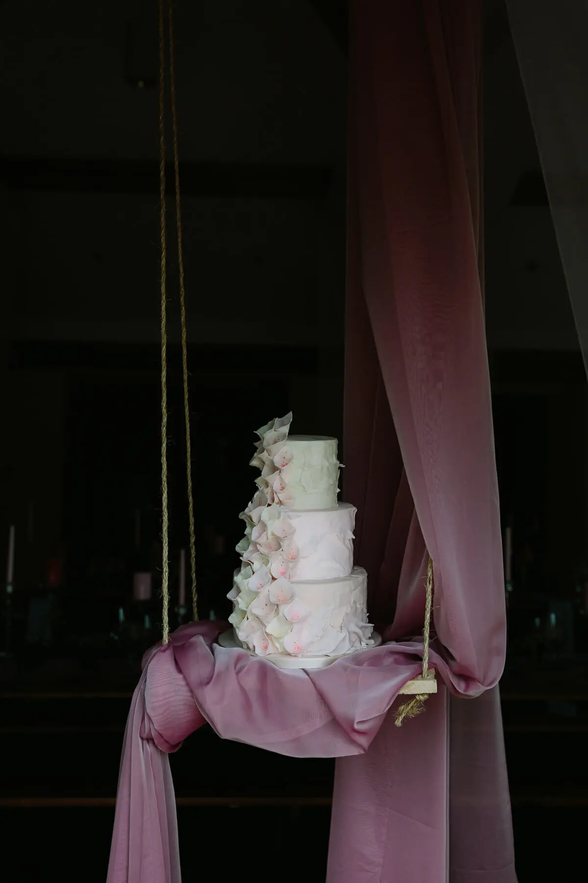 Tiered white cake with pink petal ruffles on a rope swing draped in pink fabric at Sulgrave Manor