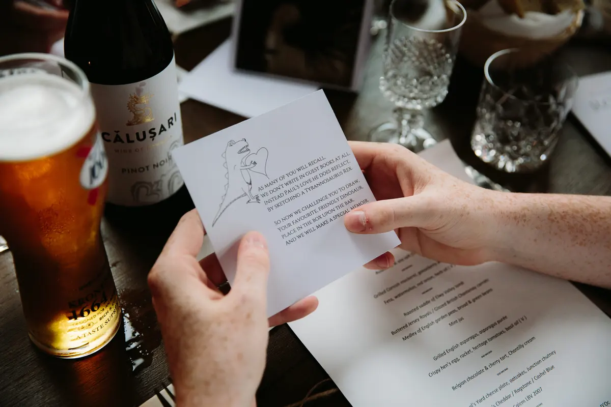 Guest holding a card with a dinosaur sketch over a menu, beside a pint of beer and a bottle of Pinot Noir