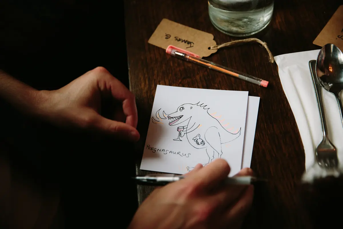 Guest drawing a cartoon dinosaur on a card at a wooden table set with a name tag and cutlery