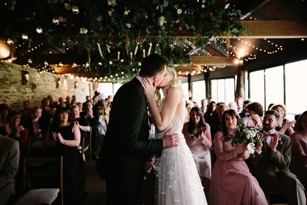 Bride and groom sharing their first kiss as guests clap beneath a canopy of greenery and fairy lights