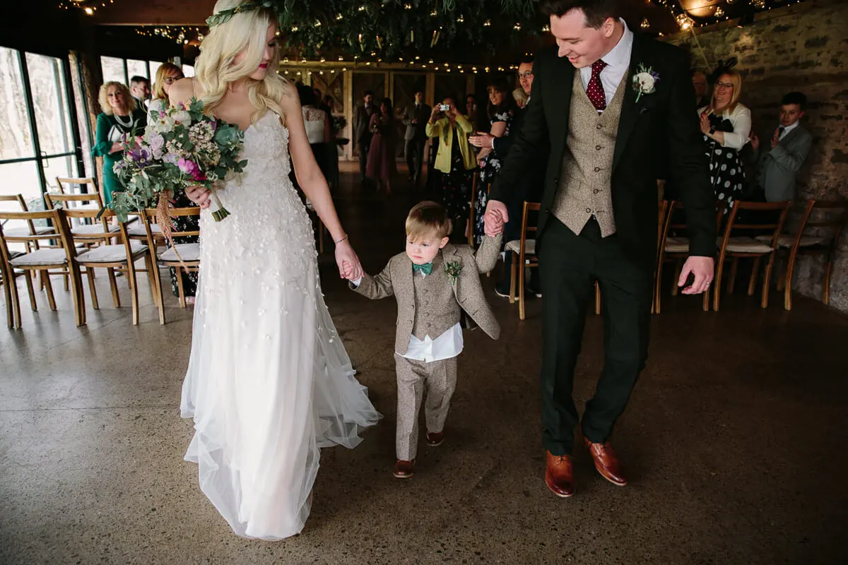 Bride and groom walking back up the aisle holding hands with a page boy between them
