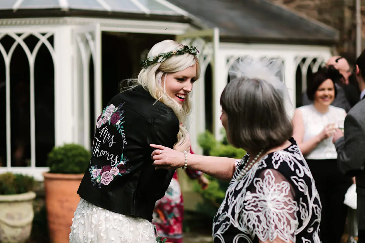 Bride in a hand-painted personalised leather jacket and flower crown greeting a guest by a conservatory