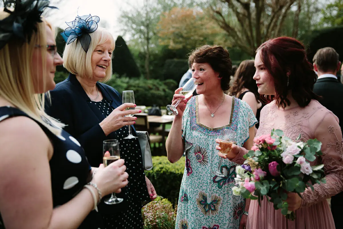 Wedding guests chatting with glasses of fizz in a garden, one bridesmaid holding a pink bouquet