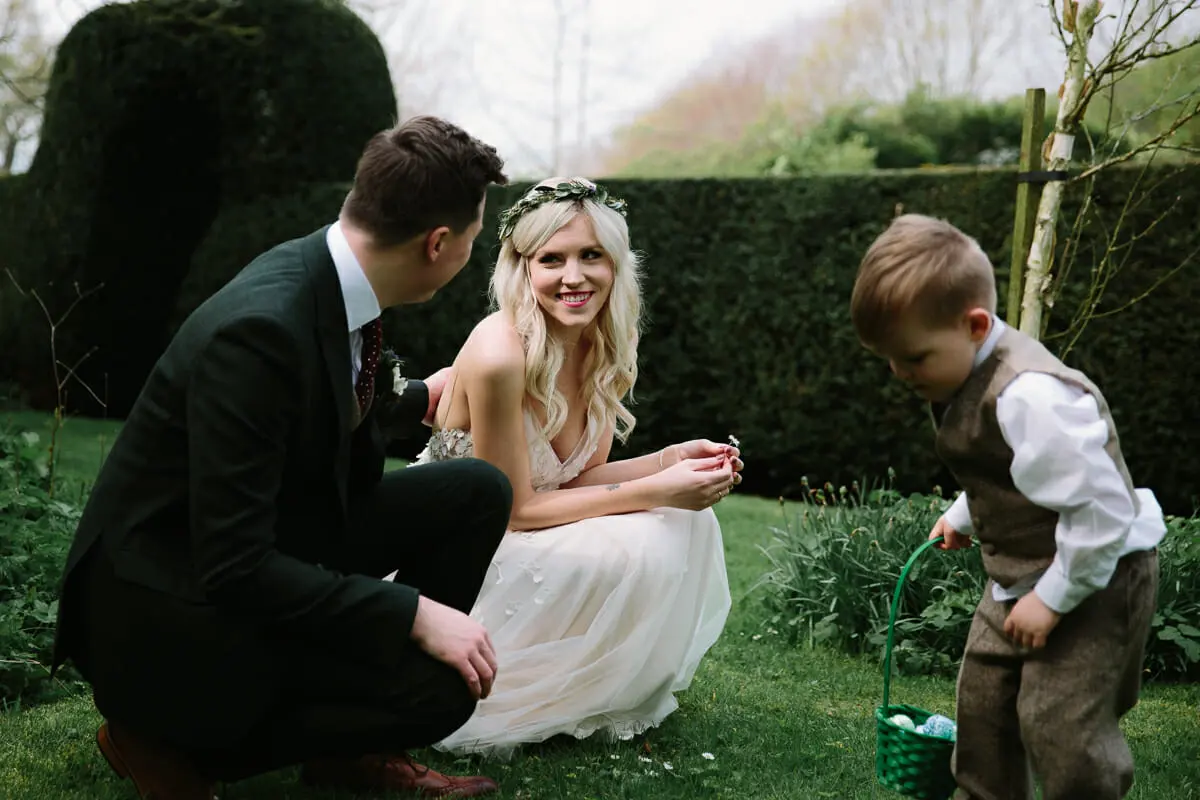 Bride and groom crouching on the lawn as a page boy with a green basket hunts for eggs by a yew hedge