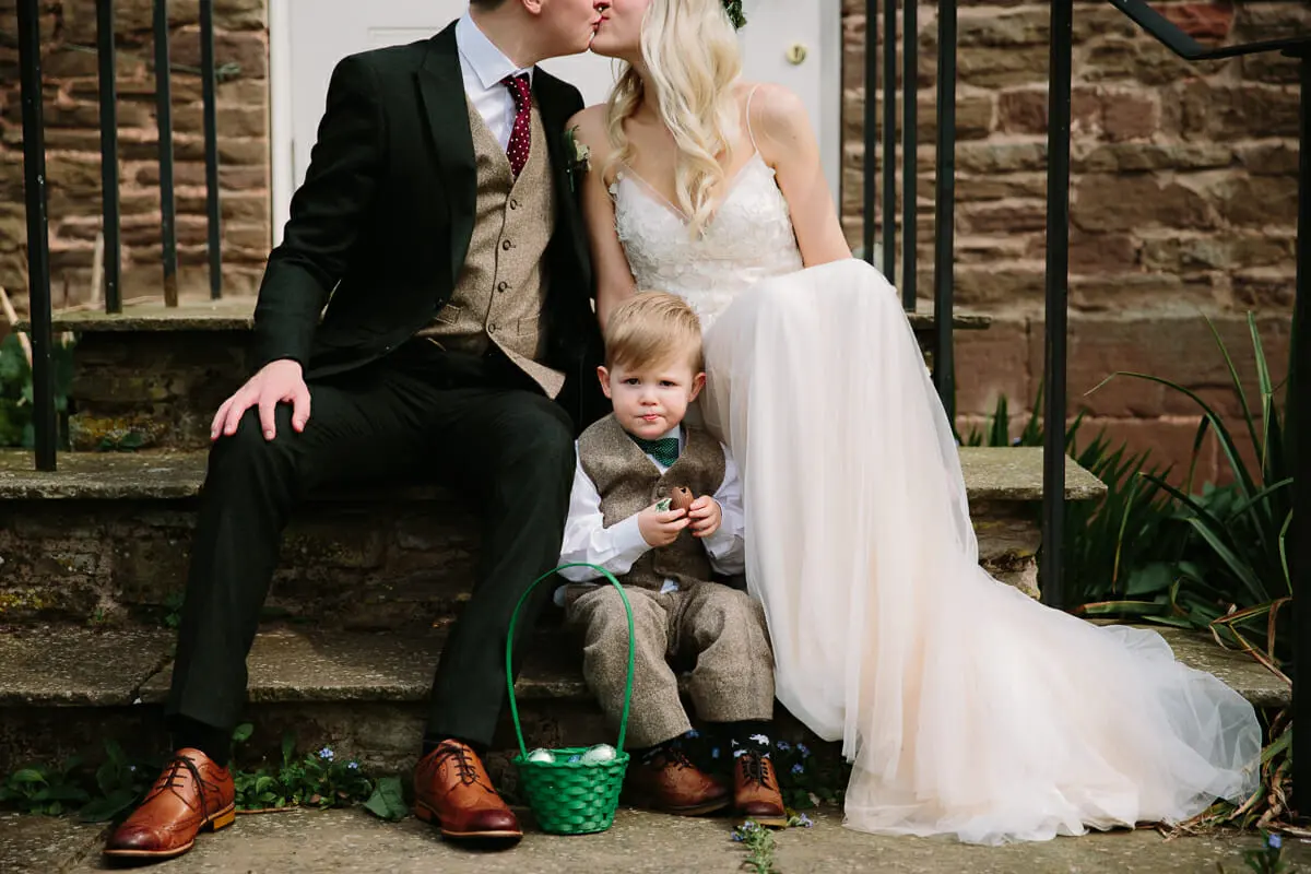 Bride and groom kissing on stone steps with a page boy sitting between them holding a green basket