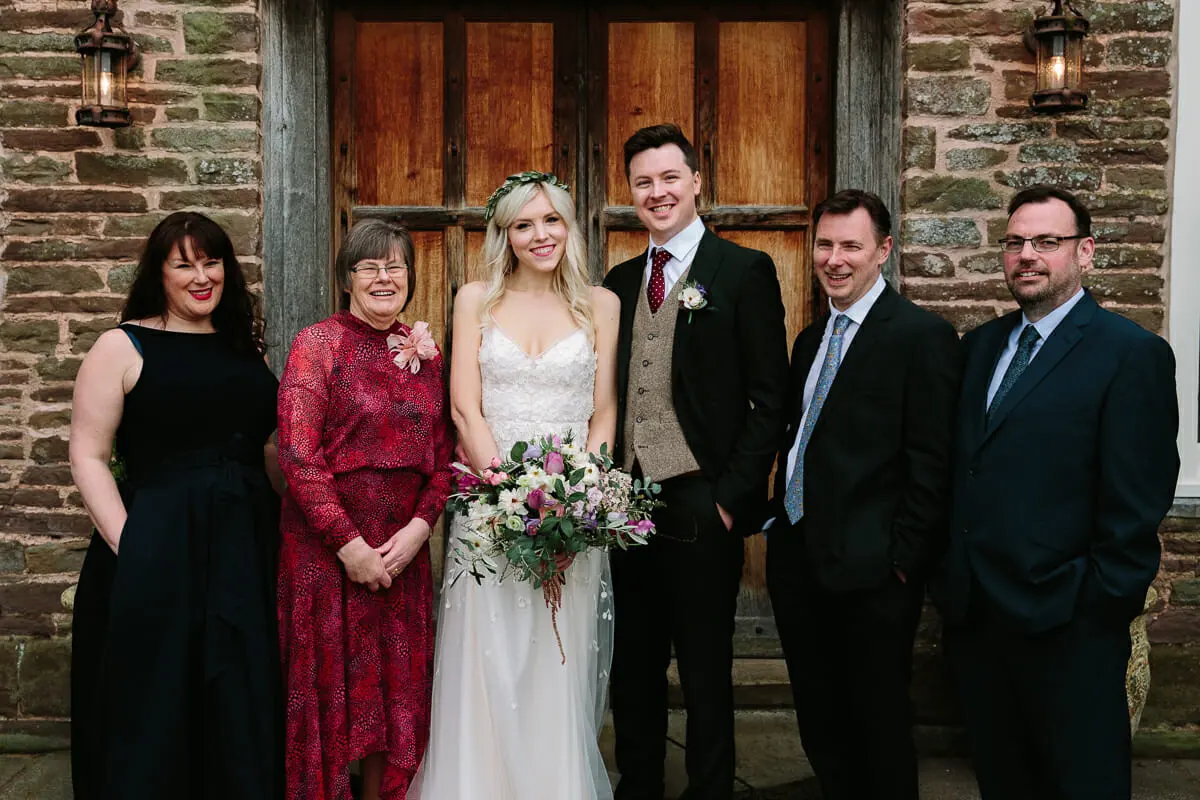Bride and groom in a family group photo outside the wooden doors of a stone building