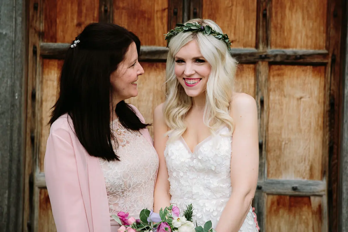 Bride in a leafy crown smiling beside a woman in a pink cardigan against old wooden doors
