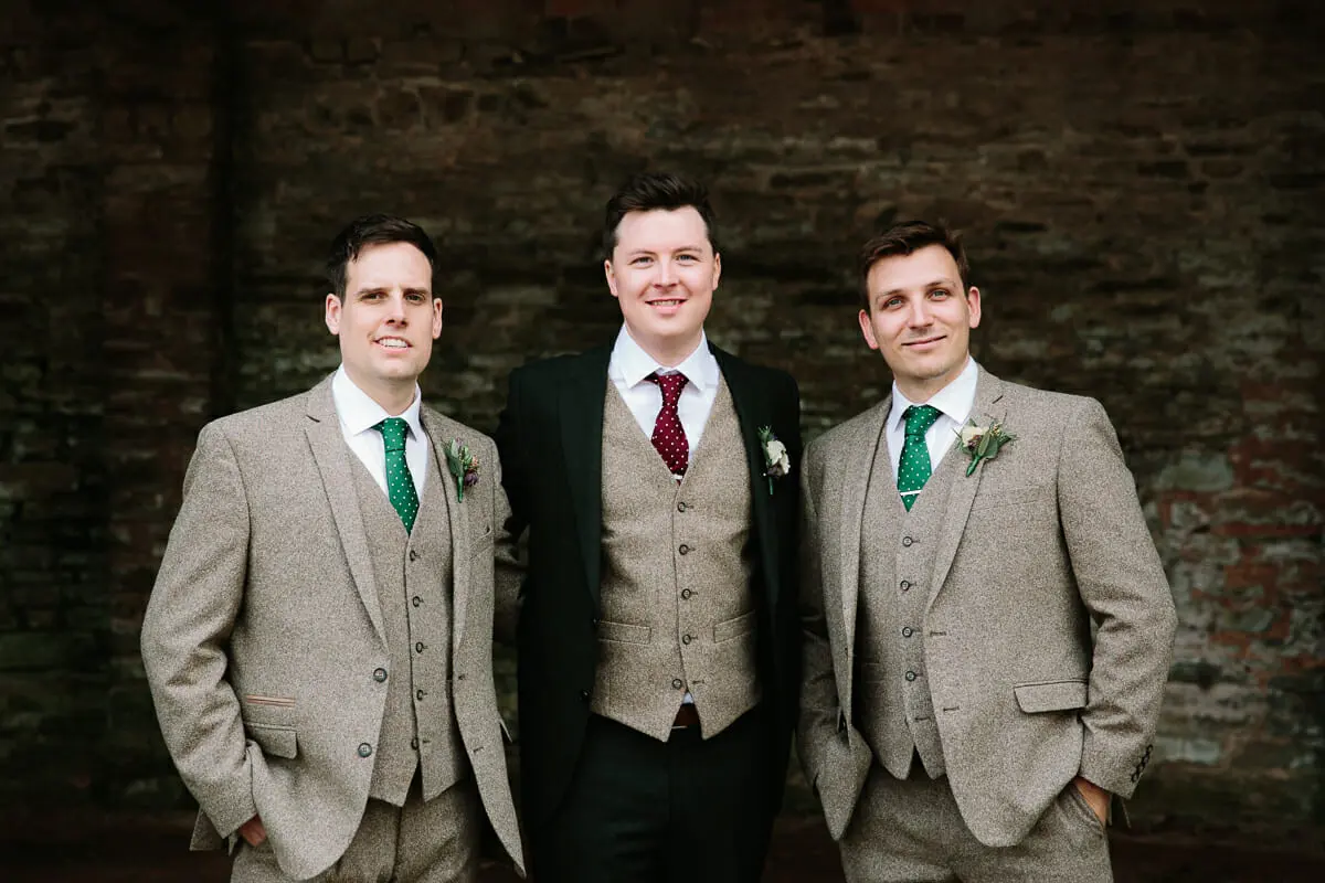 Groom in a dark suit and tweed waistcoat with two groomsmen in tweed suits against a brick wall