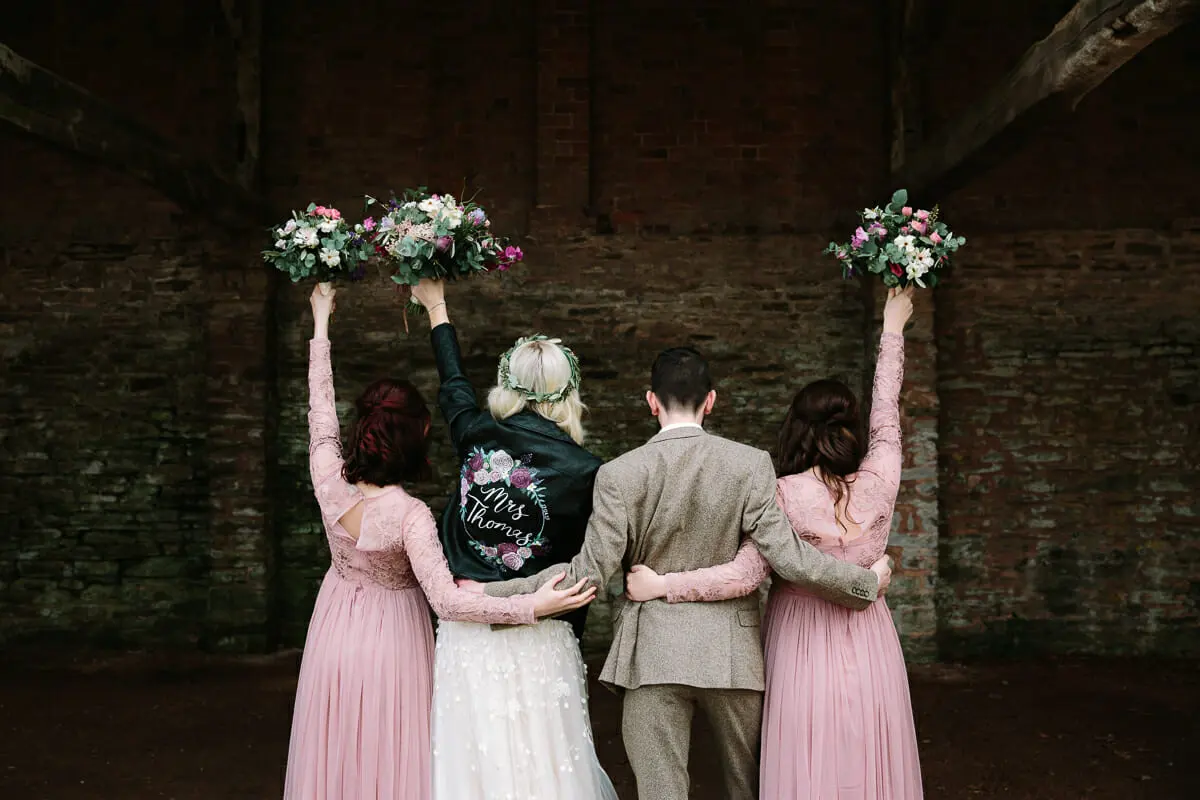 Bride, groom and bridesmaids seen from behind, arms round each other and bouquets raised high