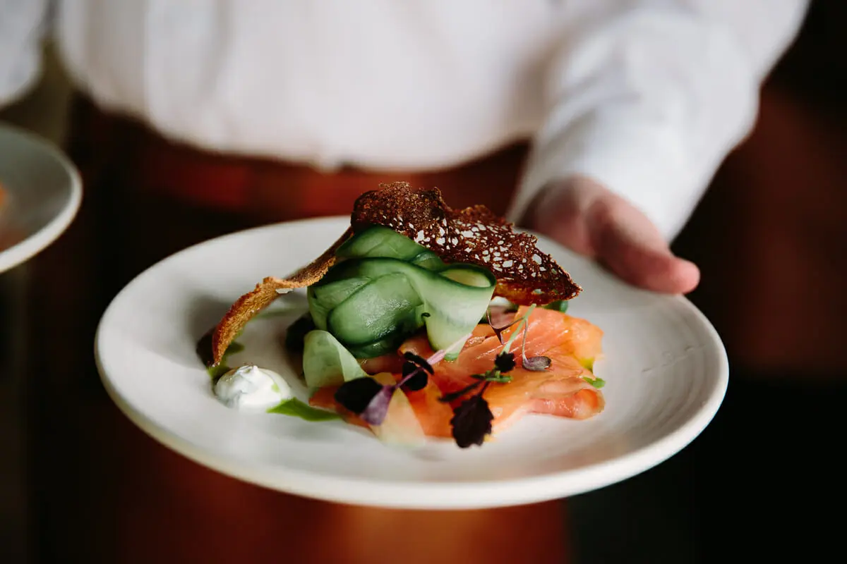 Waiter holding a plate of smoked salmon with ribbons of cucumber and a crisp rye wafer