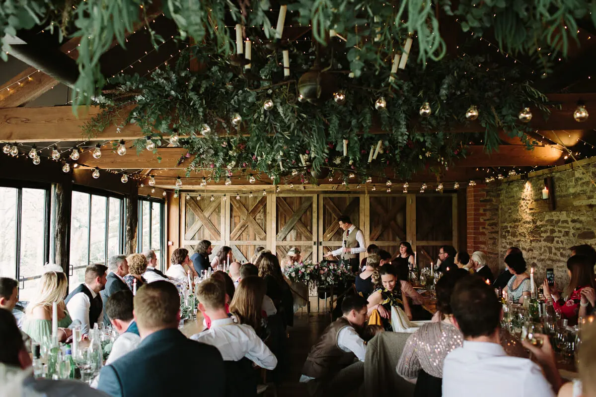 Guests listening to a speech at long tables under hanging greenery and festoon lights in a barn