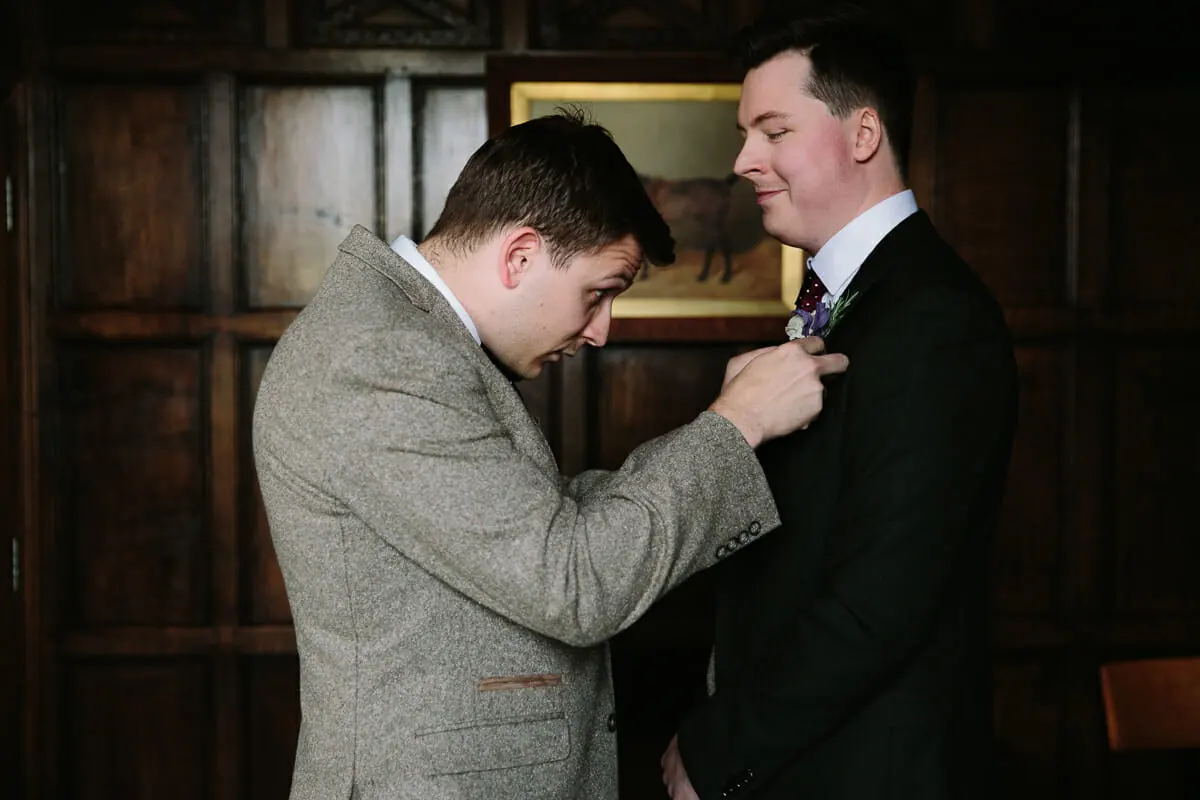 Man in a tweed jacket pinning a buttonhole on the groom in a wood-panelled room