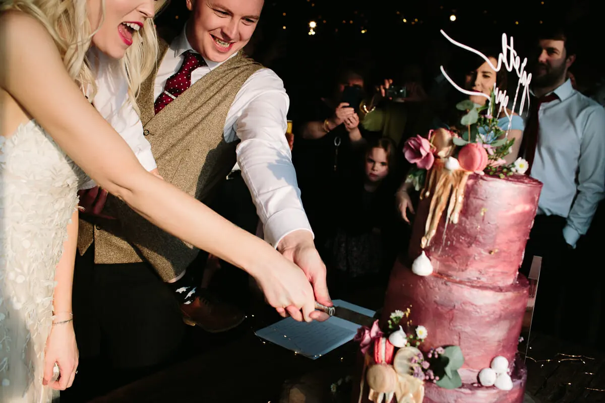 Bride and groom cutting a tall pink iced wedding cake as guests watch