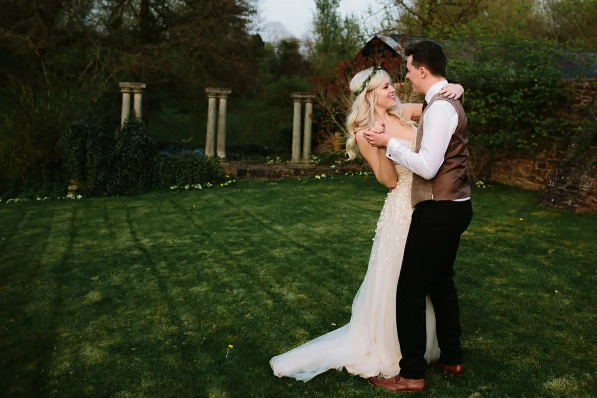Bride and groom embracing on a lawn in a walled garden with stone columns behind