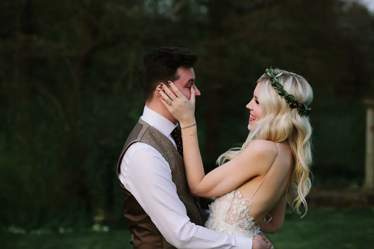 Bride in a leafy crown cupping the groom's face as he wipes away a tear