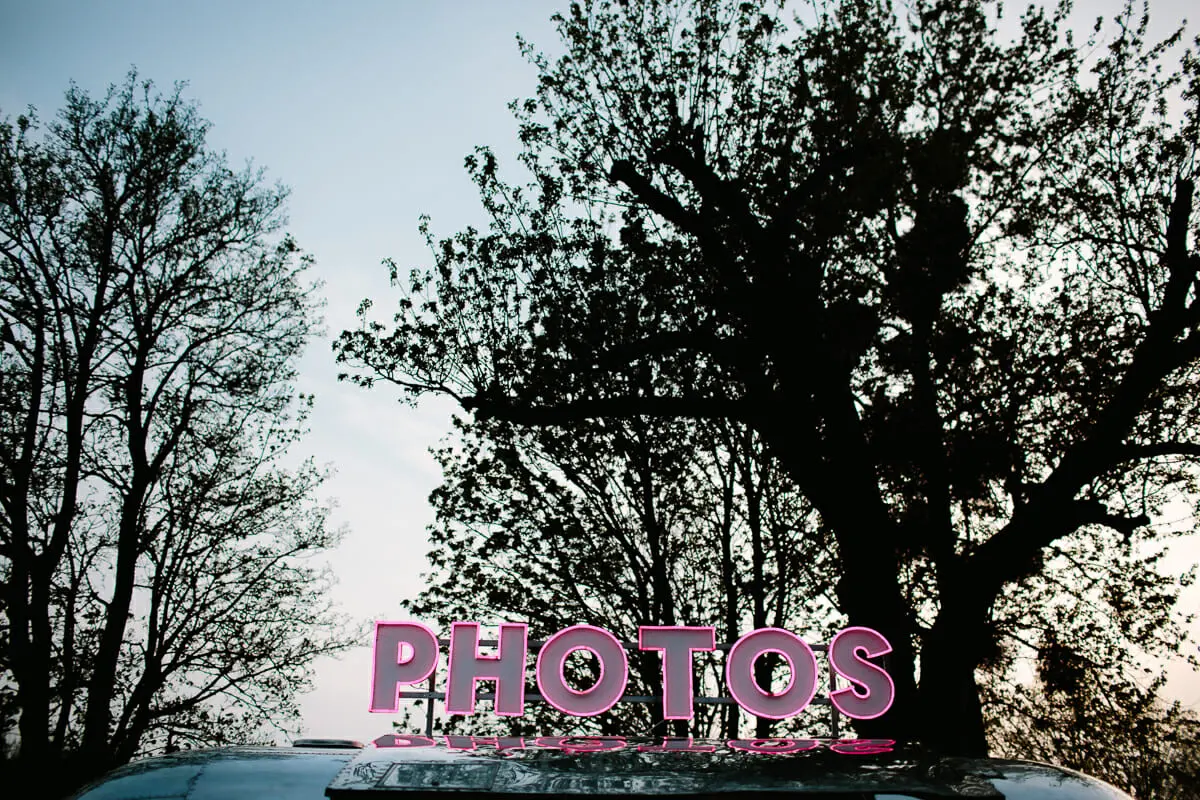 Illuminated pink PHOTOS sign on top of a photo booth van against silhouetted trees at dusk