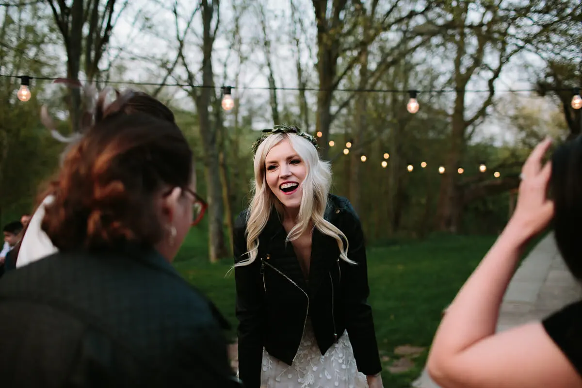 Bride in a black leather jacket laughing with guests under festoon lights in the evening