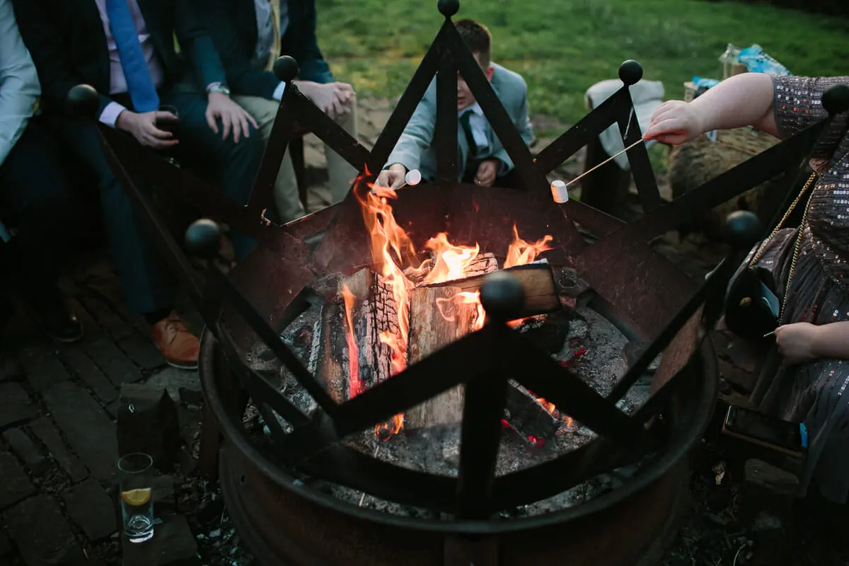 Guests toasting marshmallows over a fire pit at an evening wedding reception