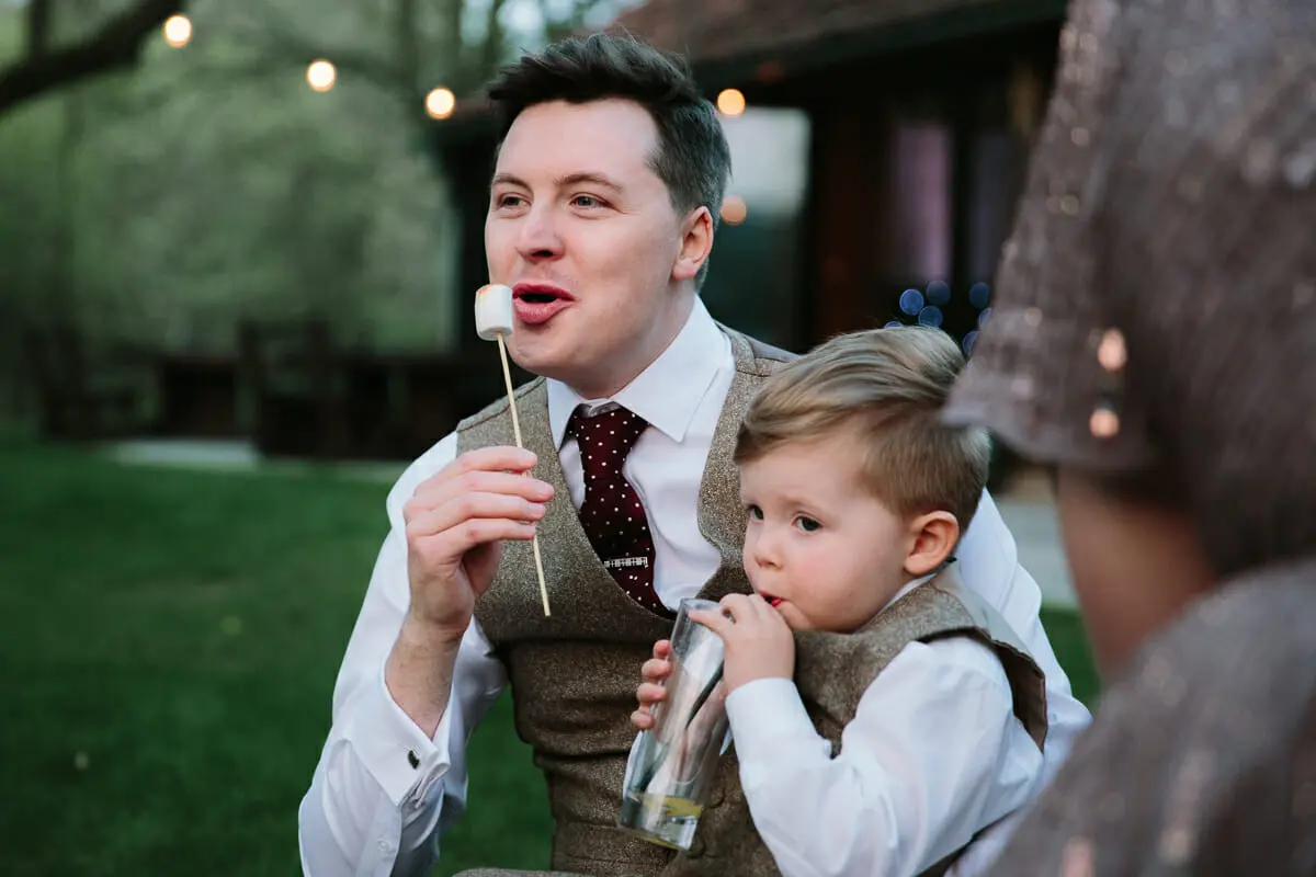 Groom eating a toasted marshmallow while holding a toddler in a matching tweed waistcoat