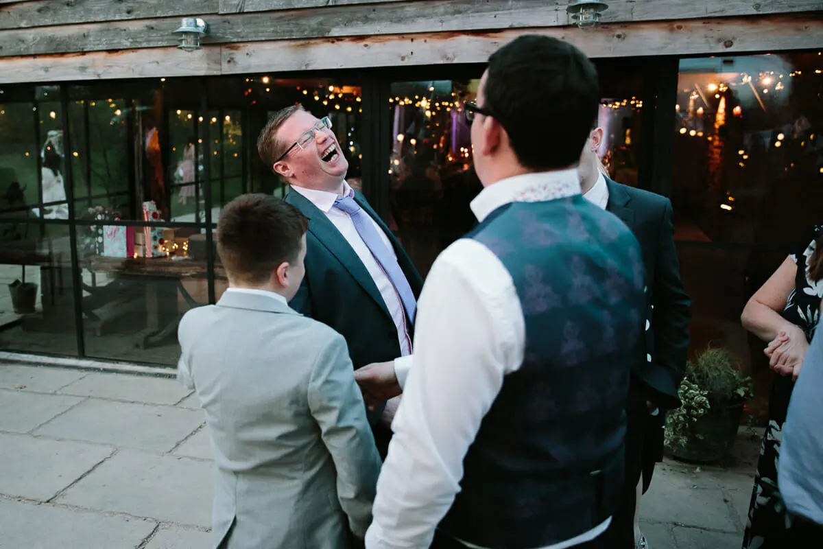 Wedding guest in a suit roaring with laughter outside a barn lit with fairy lights