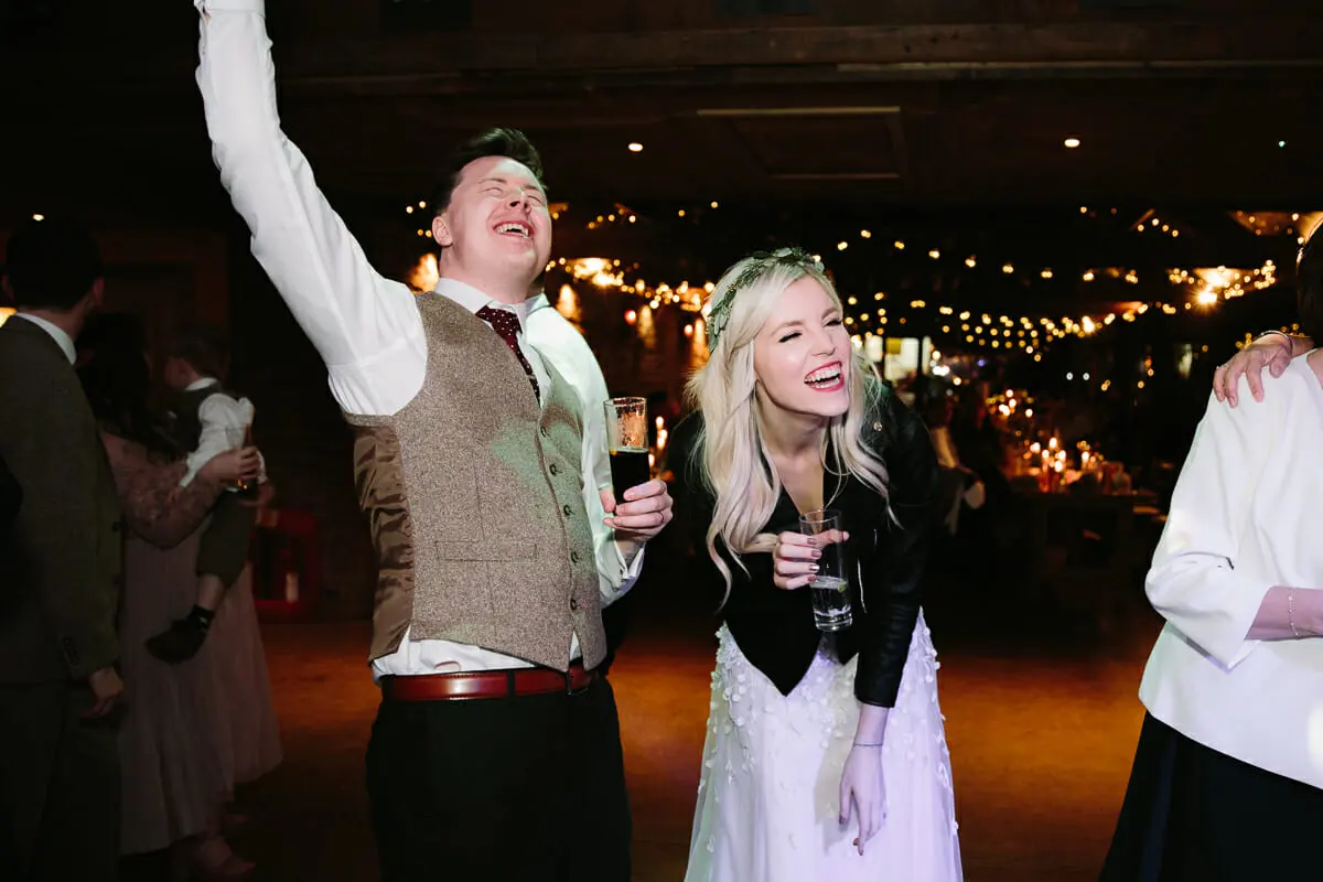 Groom throwing his arm in the air and bride laughing with drinks on the dance floor