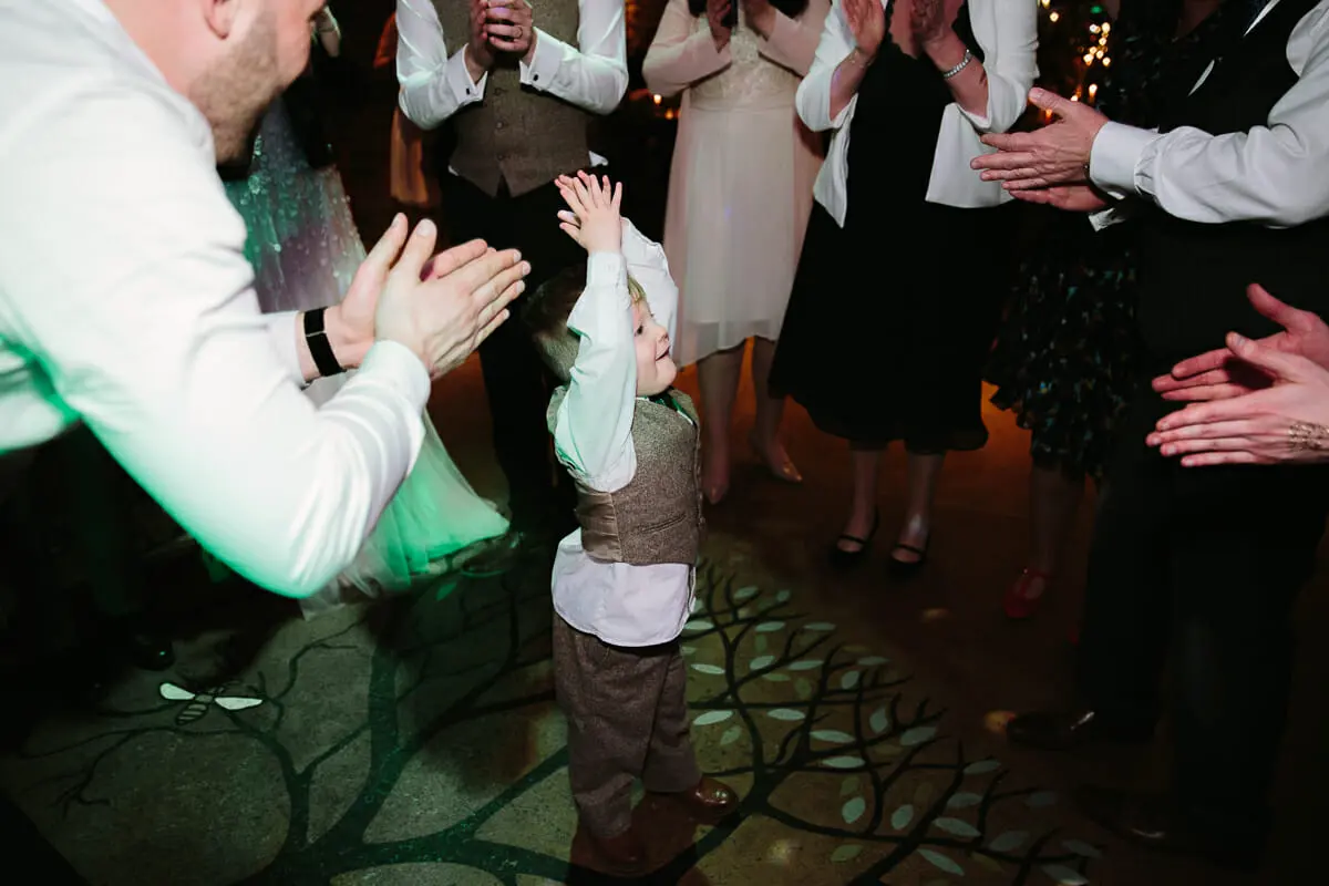 Toddler in a tweed waistcoat raising his hands on the dance floor as guests clap around him