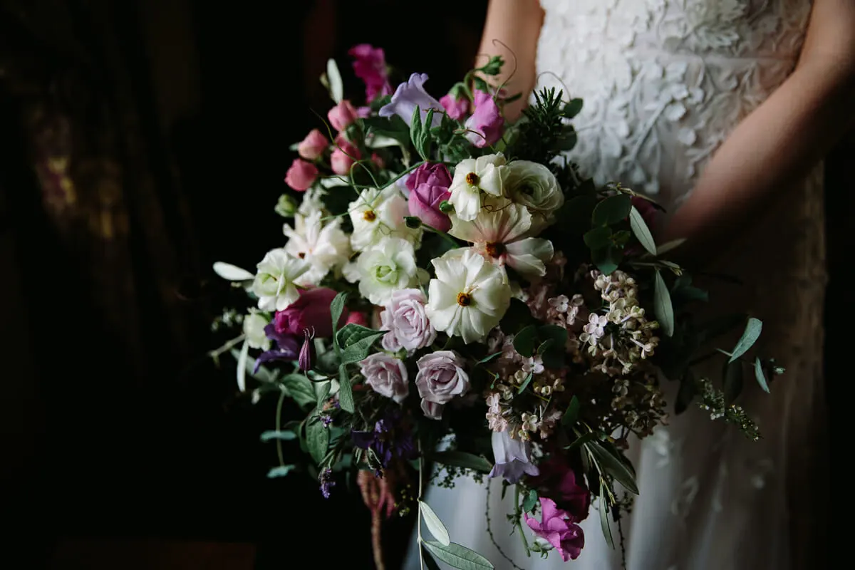Bride holding a loose bouquet of pink and lilac roses, white ranunculus and greenery against her lace dress