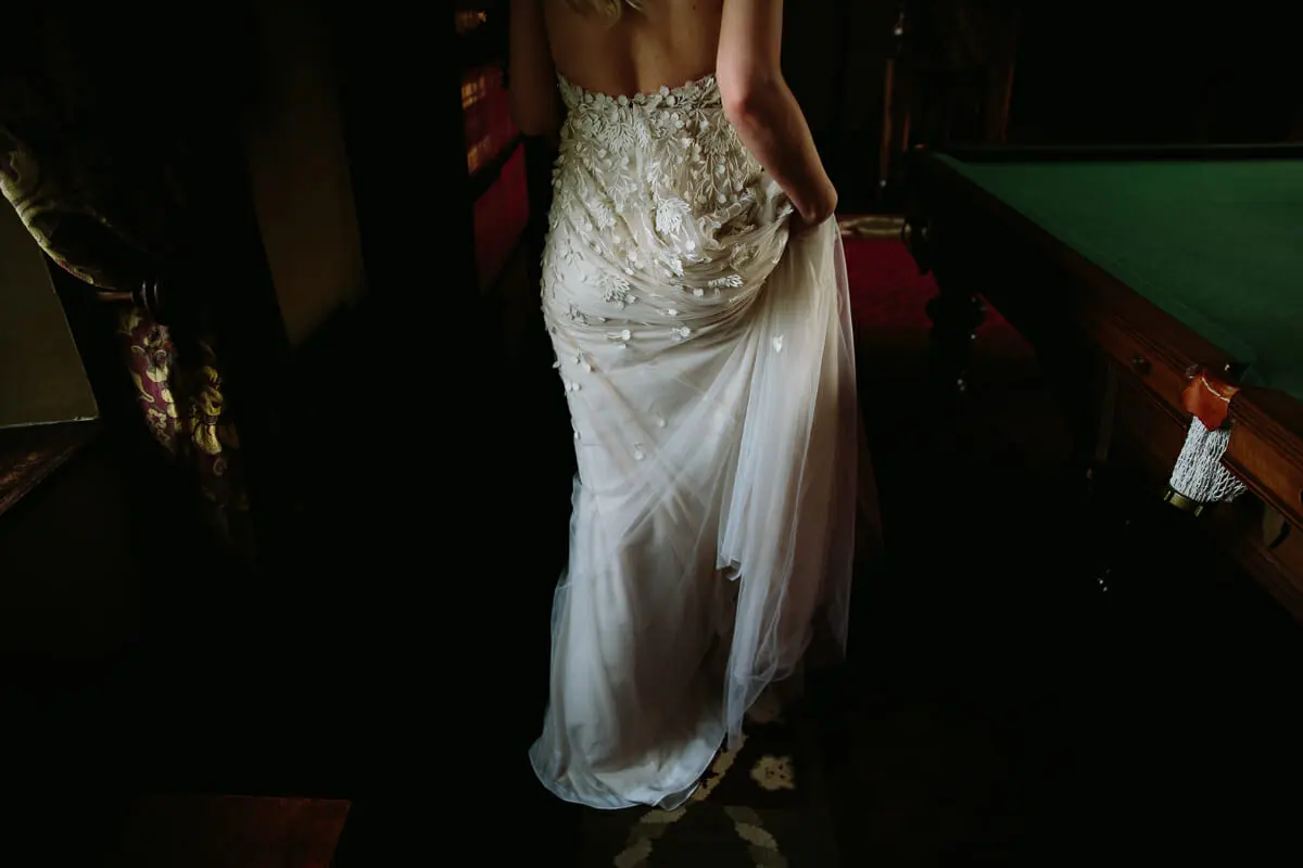 Bride lifting the train of her beaded, backless gown as she walks past a snooker table