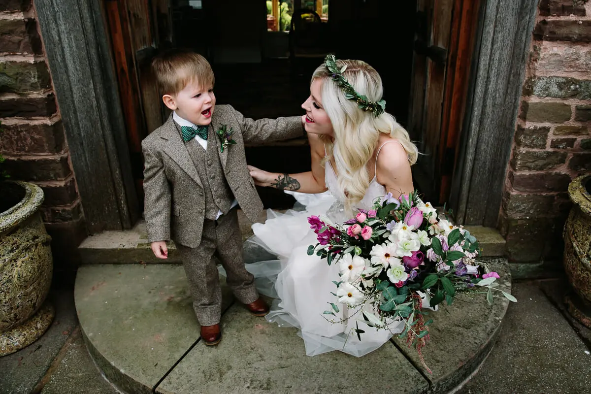 Bride in a flower crown crouching to talk to a page boy in a tweed suit on a stone doorstep