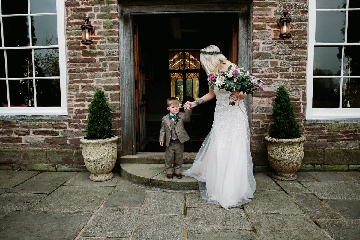 Bride holding hands with a page boy in a tweed suit at the stone doorway of a country house