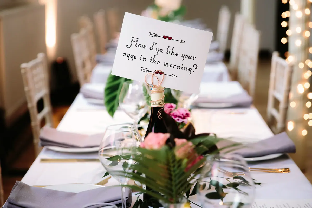 Long table with a song lyric sign on a wine bottle, pink roses, fern leaves and fairy lights behind