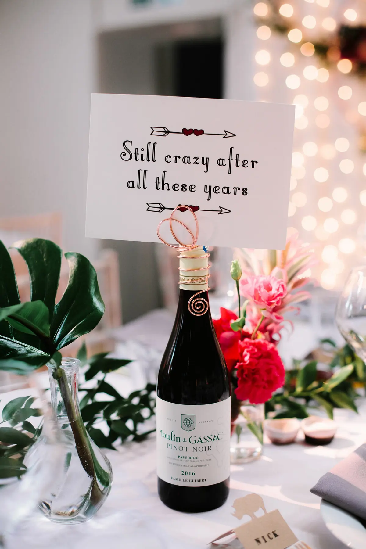 Wine bottle holding a card reading Still crazy after all these years, beside red and pink flowers and a fern leaf