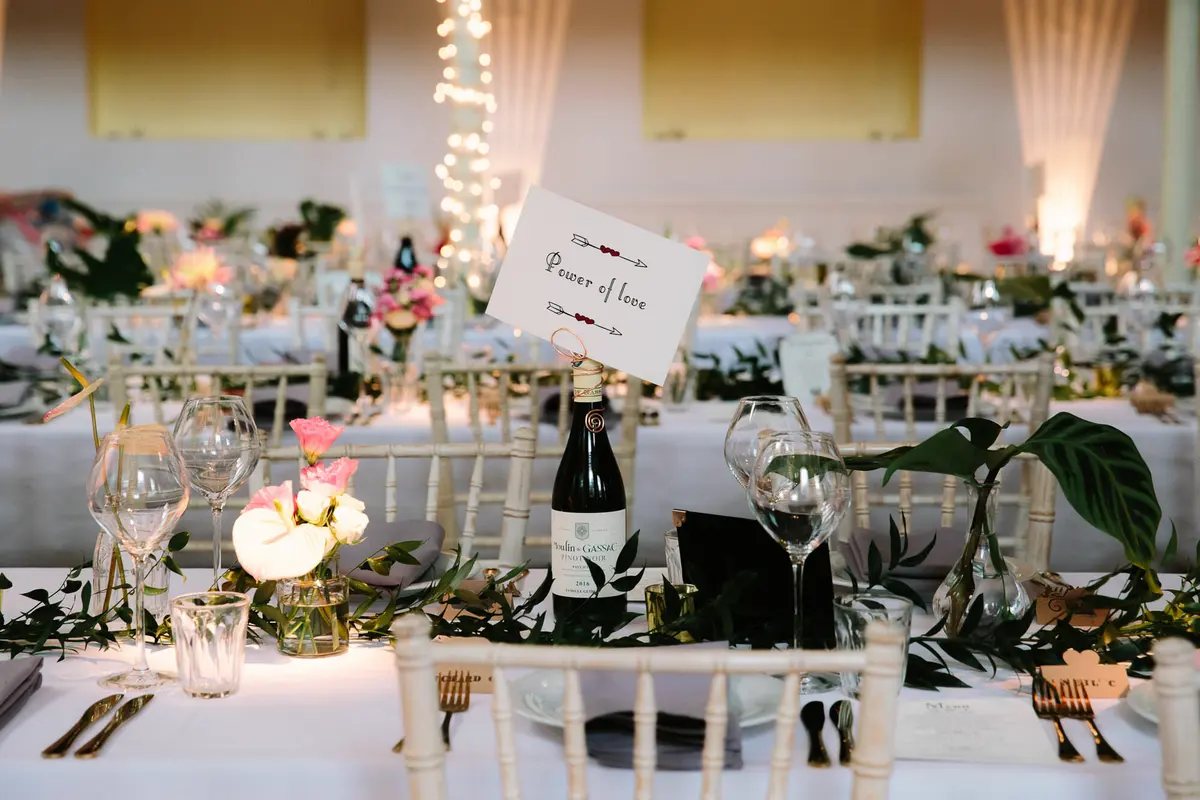 Reception tables with white chairs, wine bottles holding song title cards, pink flowers and greenery