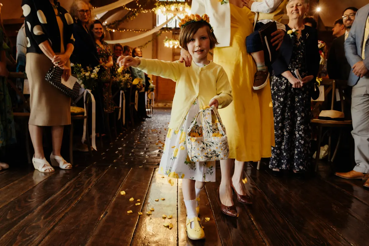Flower girl in a yellow cardigan scattering petals down the aisle of a barn ceremony
