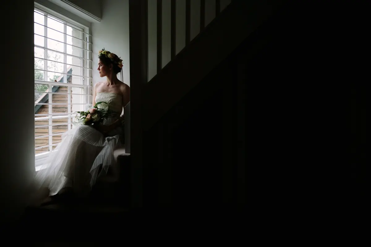 Bride in a flower crown sitting on the stairs by a window, holding her bouquet