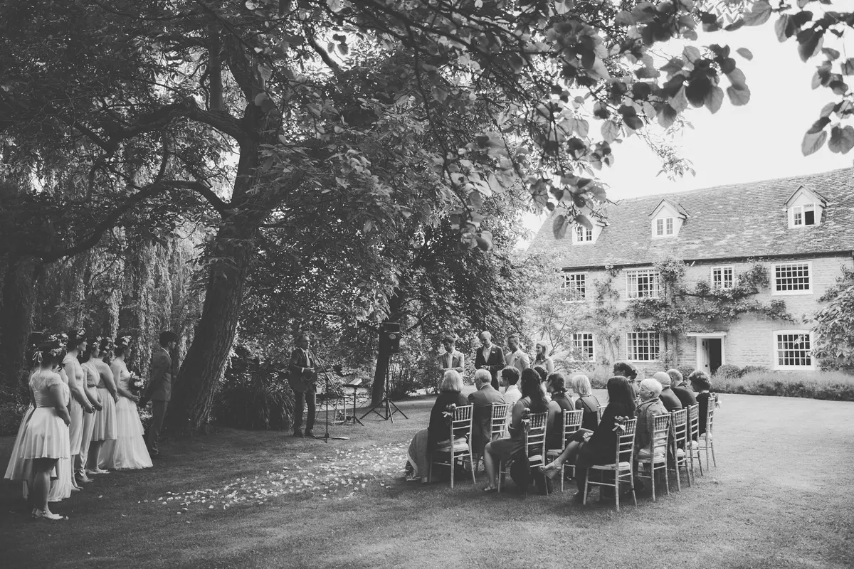 Black and white view of an outdoor ceremony under a large tree beside a stone country house