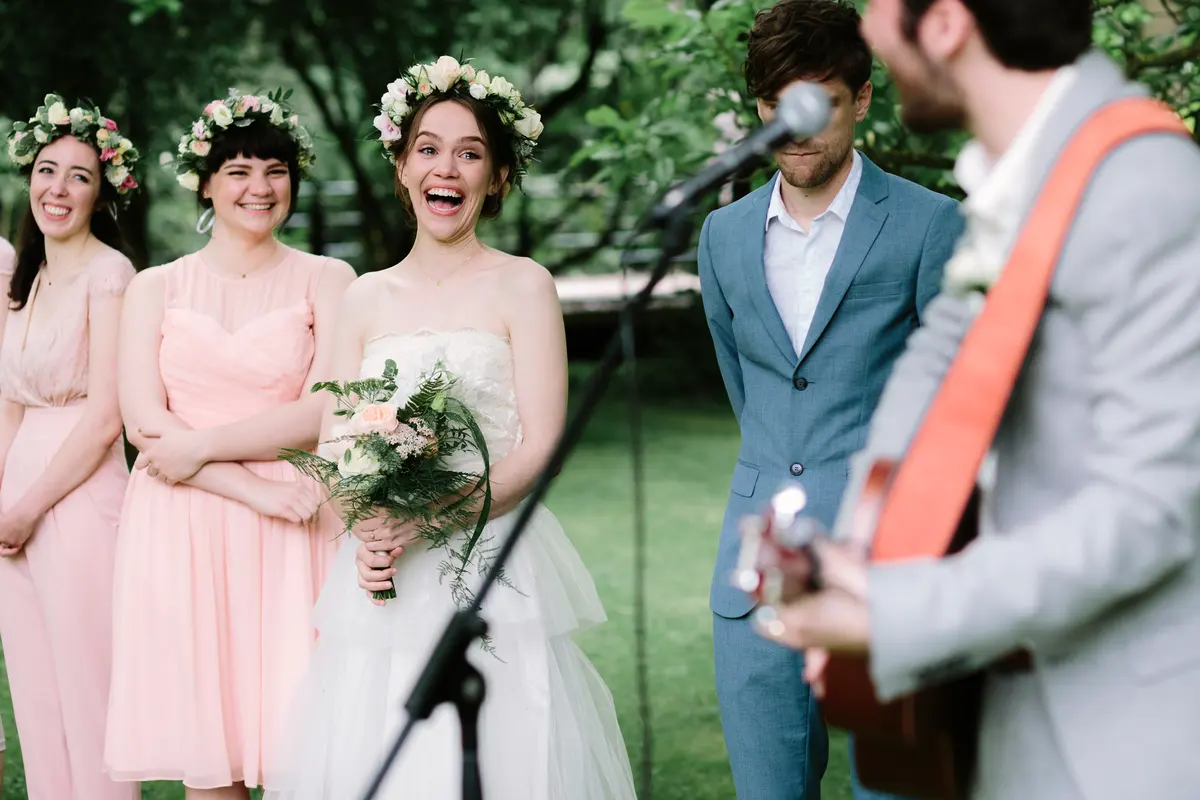 Bride in a flower crown laughing as a guitarist sings during the outdoor ceremony