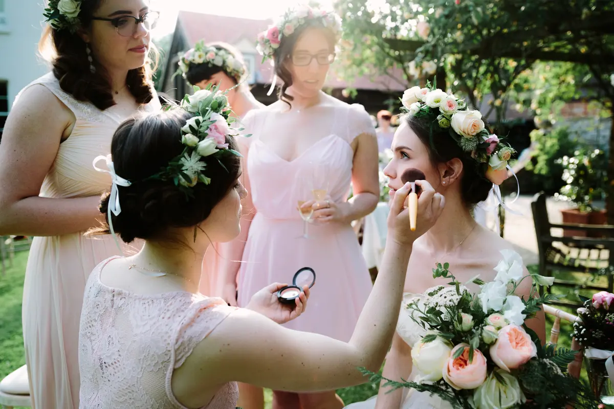 Bridesmaid touching up the bride's make-up with a brush in the sunny garden