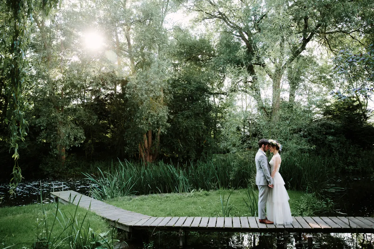Bride and groom embracing on a wooden boardwalk by a pond surrounded by trees