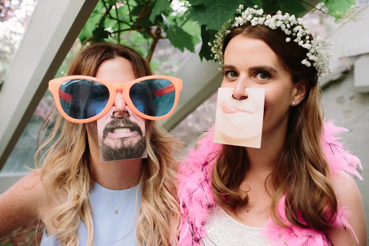 Bride in a flower crown and a friend posing with paper face props and giant sunglasses