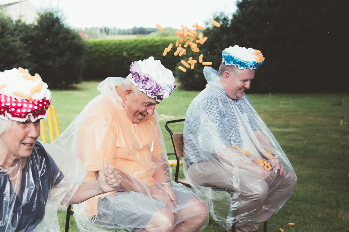 Guests in shower caps and plastic ponchos laughing as cheese puffs are thrown at them on a lawn