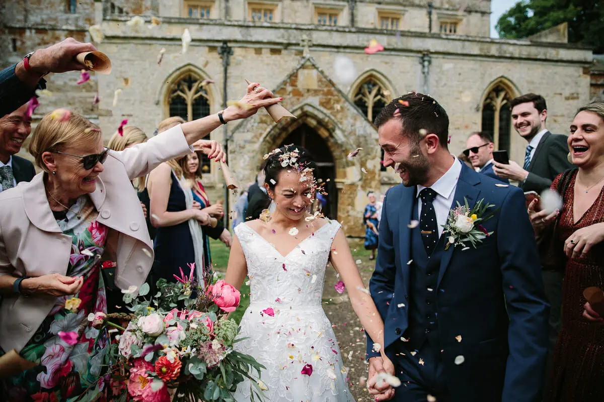 Bride and groom laughing as guests shower them with confetti outside a stone church