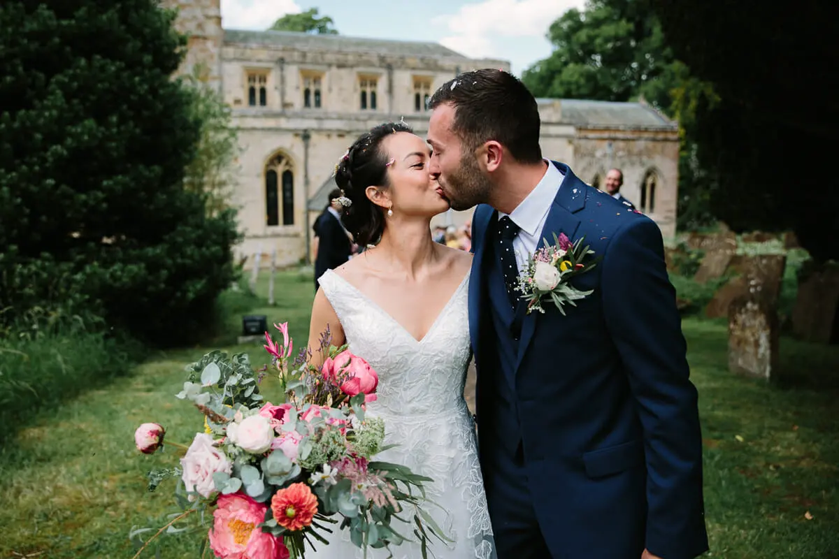 Bride and groom kissing in the churchyard, her bouquet of peonies and roses in front