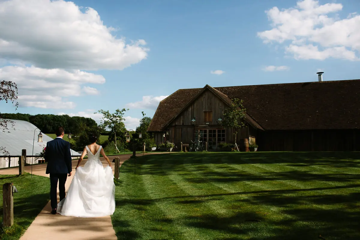 Bride and groom walking along a path towards the timber barn at Soho Farmhouse
