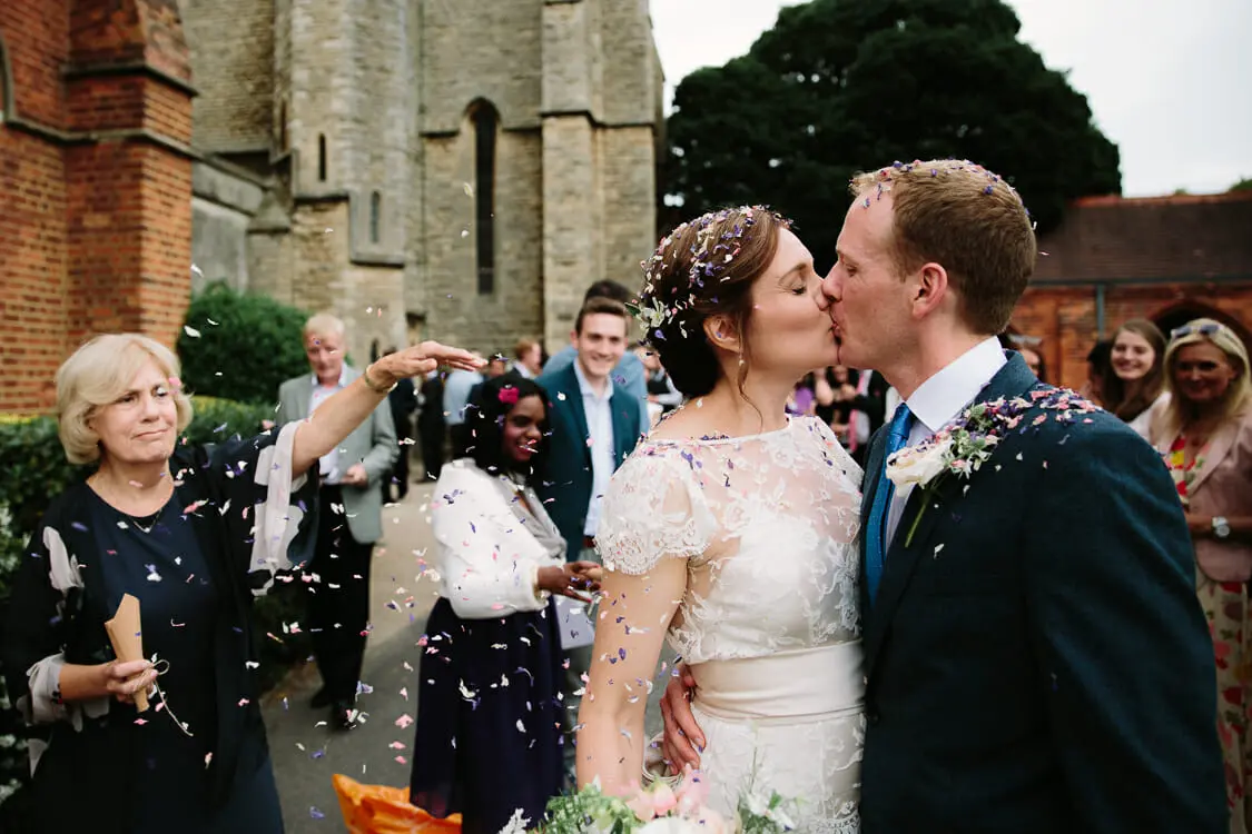 Bride and groom kissing as guests throw petal confetti outside a stone chapel