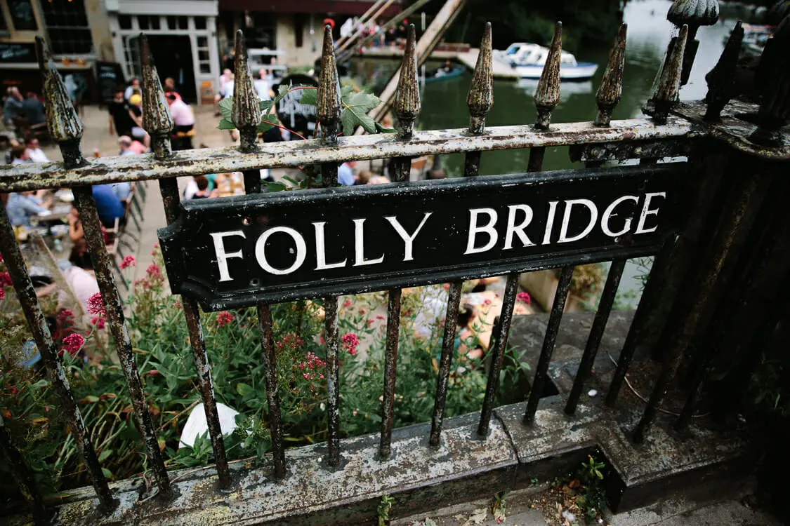 Folly Bridge sign on old iron railings above riverside drinkers and boats in Oxford