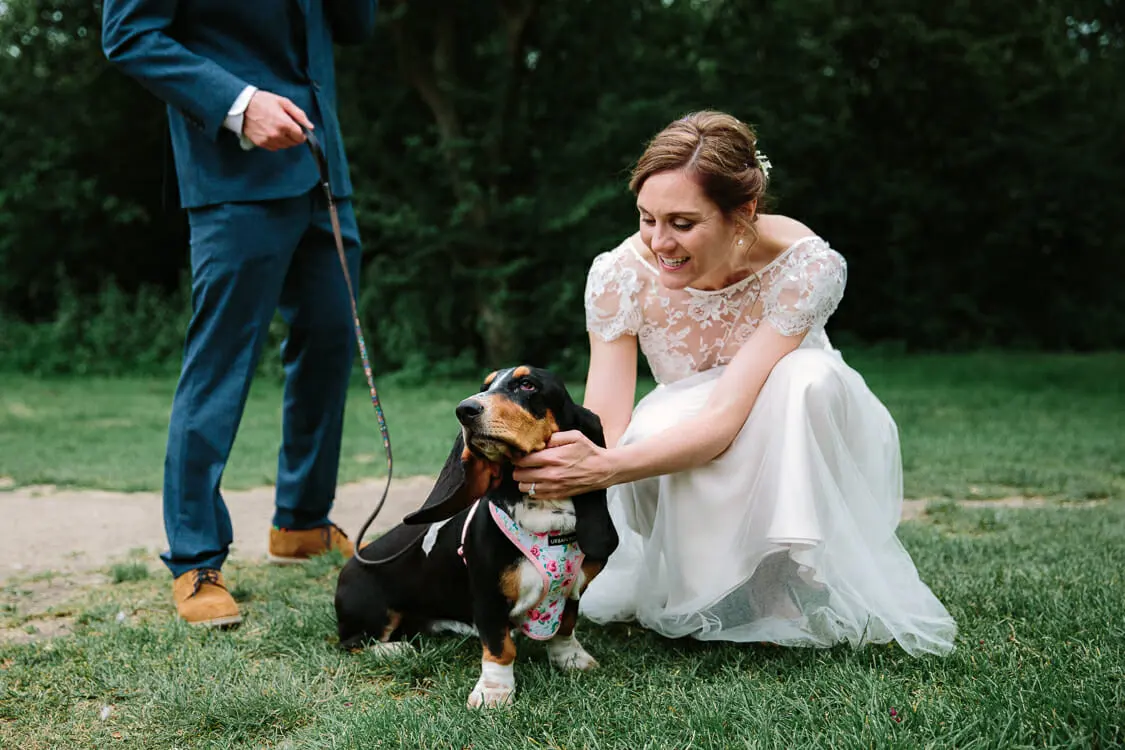 Bride crouching on the grass to stroke a basset hound wearing a floral bandana as the groom holds its lead