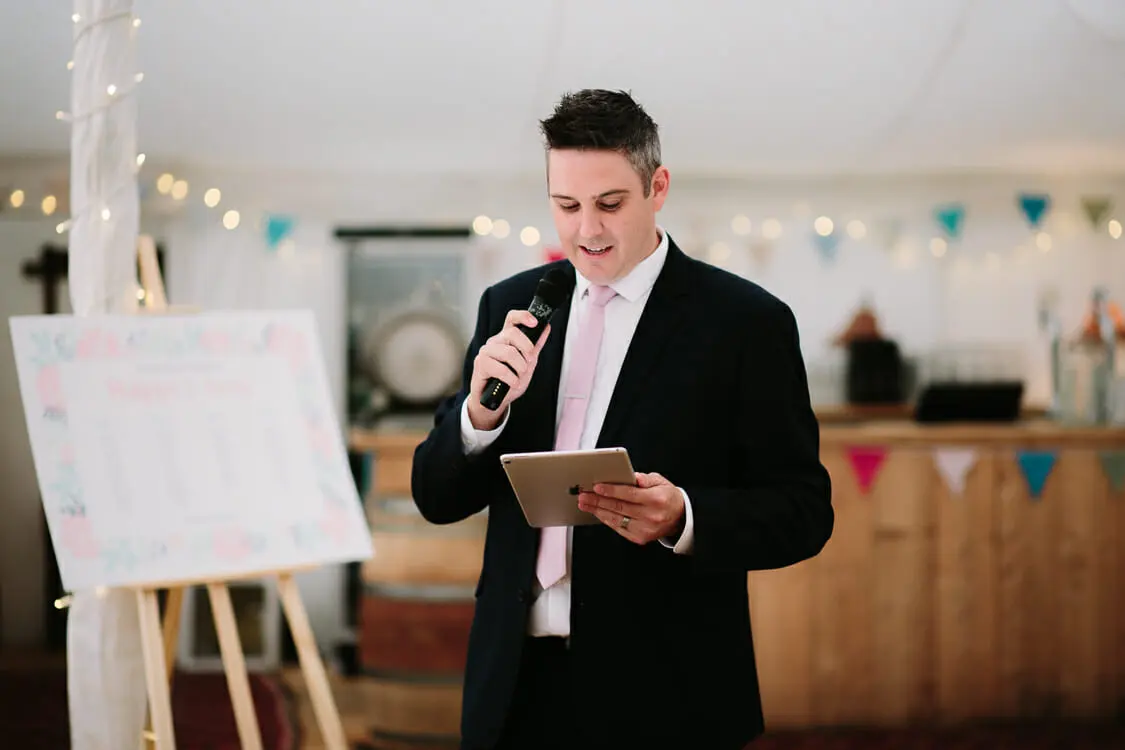 Guest giving a speech from a tablet with a microphone in a marquee hung with bunting and fairy lights