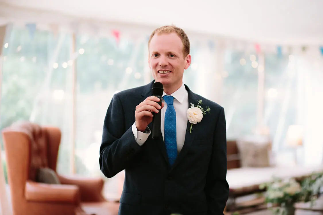 Groom smiling as he gives his speech into a microphone in the marquee at the Perch